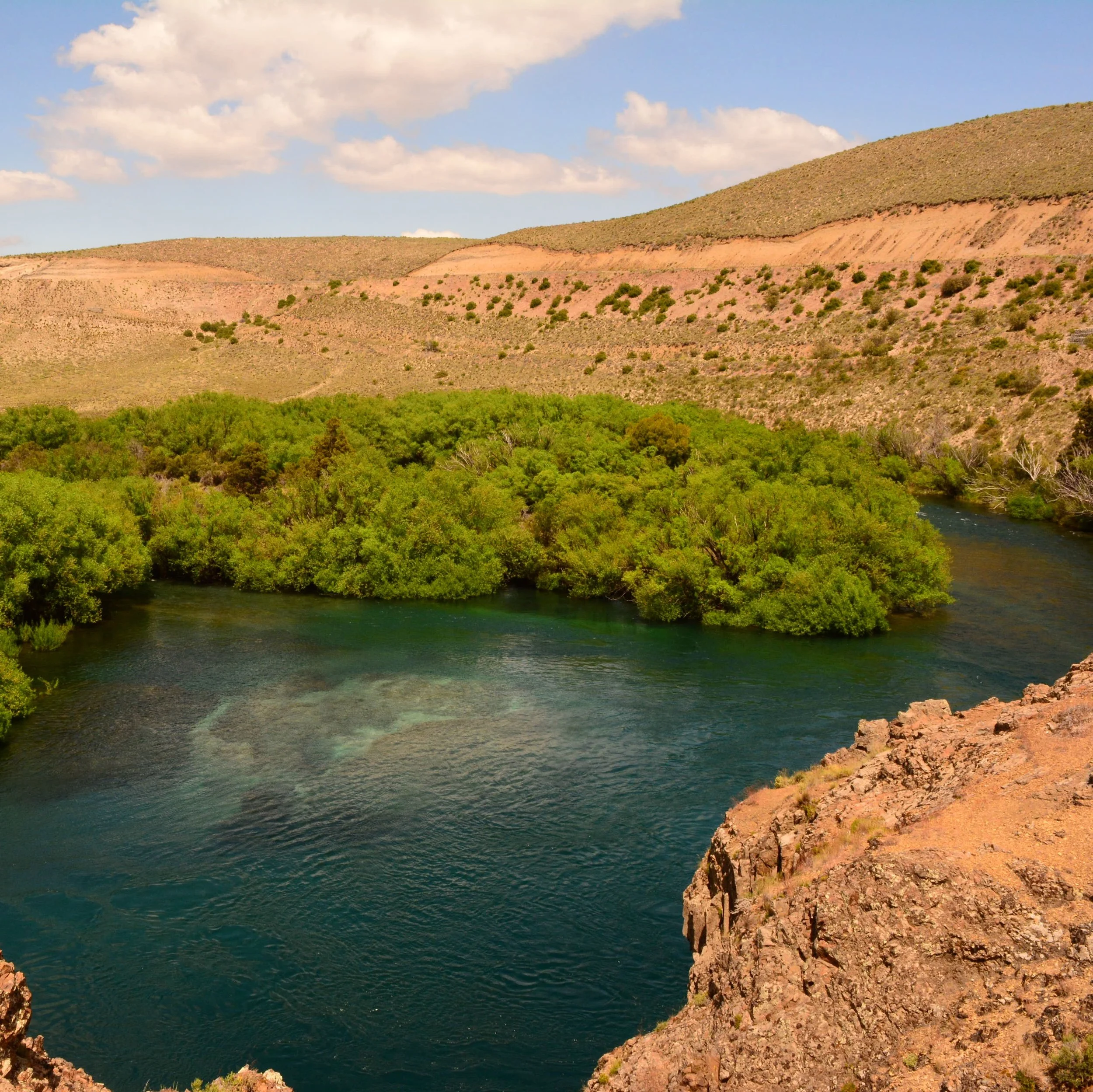 A river flowing through a lush green area with hills in the background and a partly cloudy sky.