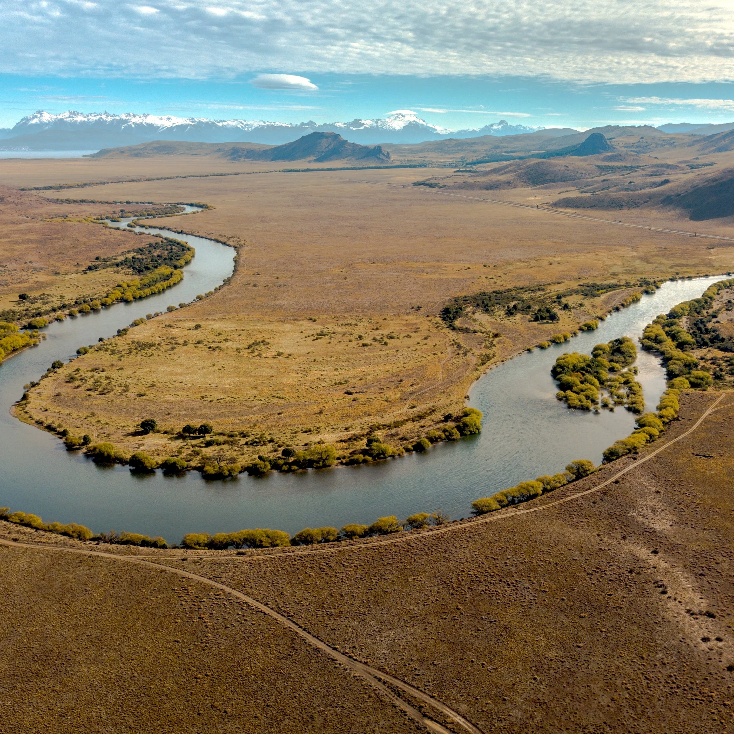 A winding river through a dry, open landscape with mountains in the background under a partly cloudy sky.