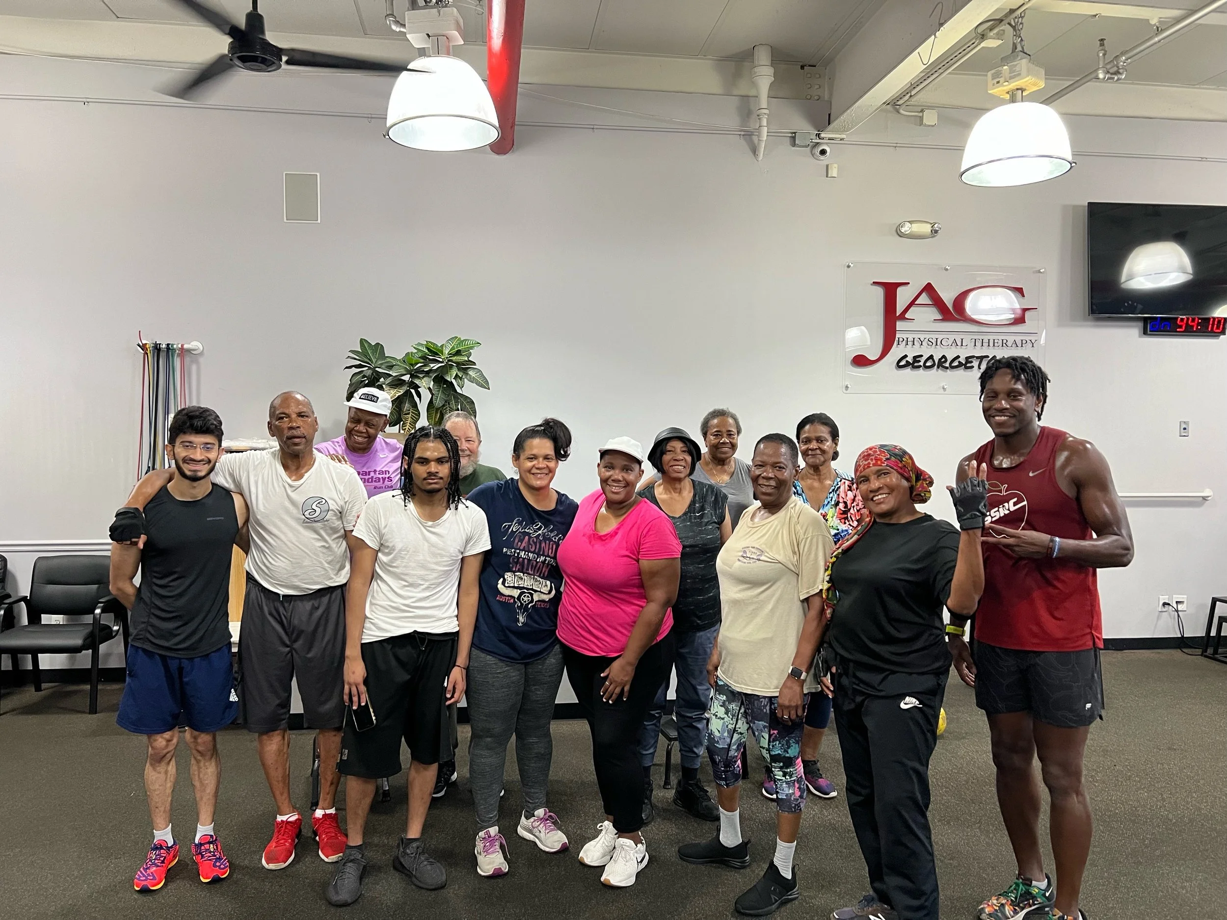 A group of diverse people posing inside a physical therapy or fitness center. They are wearing casual athletic clothing and smiling at the camera. There is a sign on the wall reading "JAC Physical Therapy Georgetown." The setting is a well-lit indoor space with exercise equipment visible.