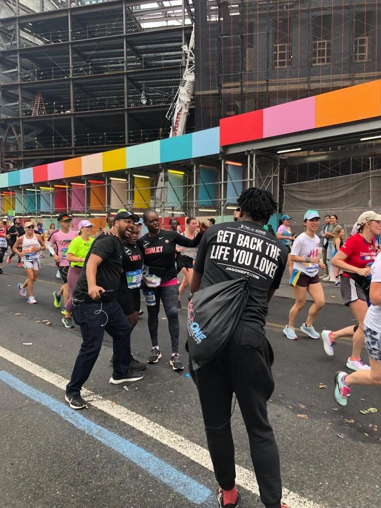 Group of runners participating in a road race, some taking a selfie, wearing athletic gear. Background includes a colorful construction site and spectators. One person wears a shirt with motivational text.