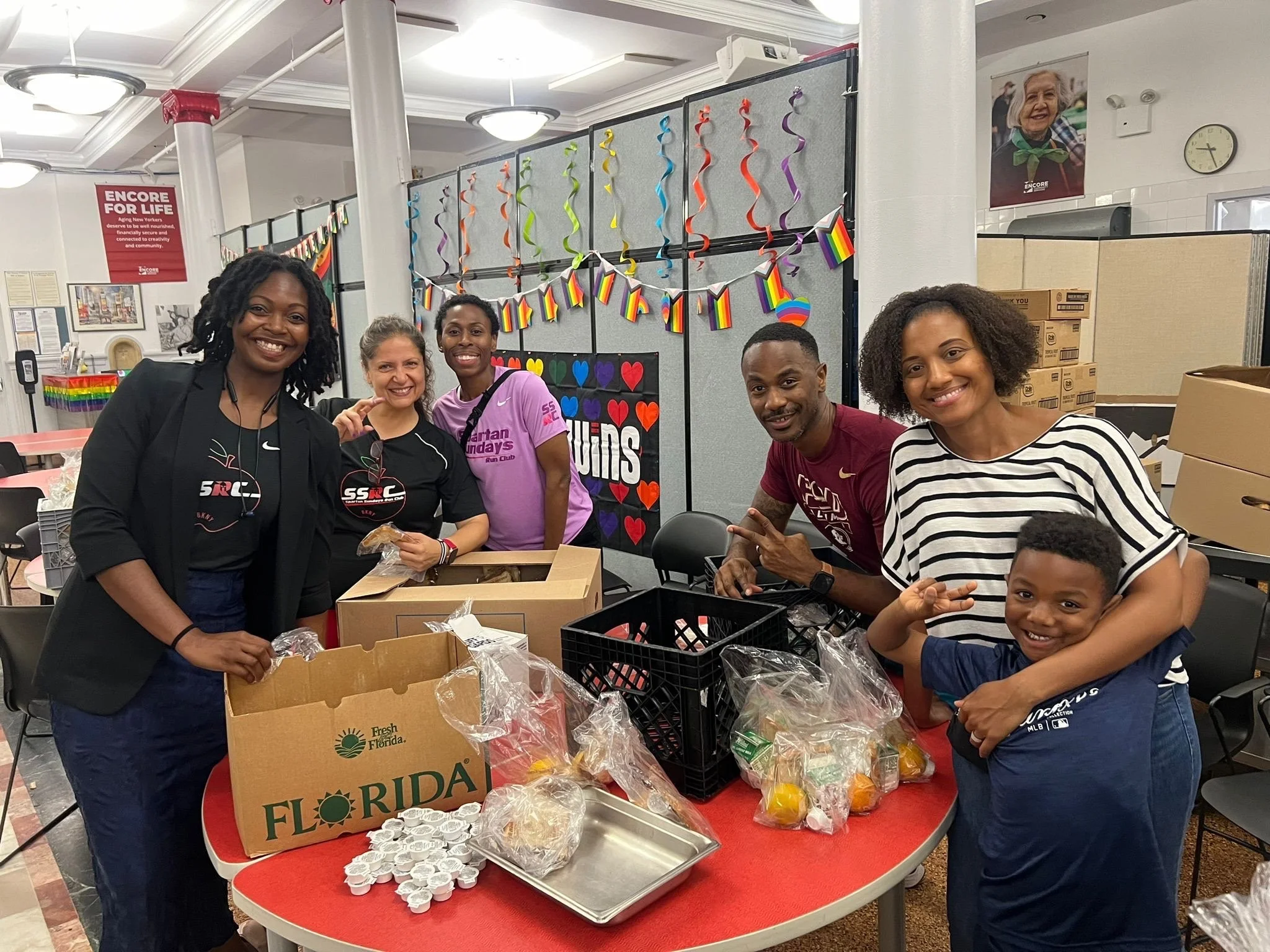 Group of people packing food in a community center with rainbow and heart decorations in the background.