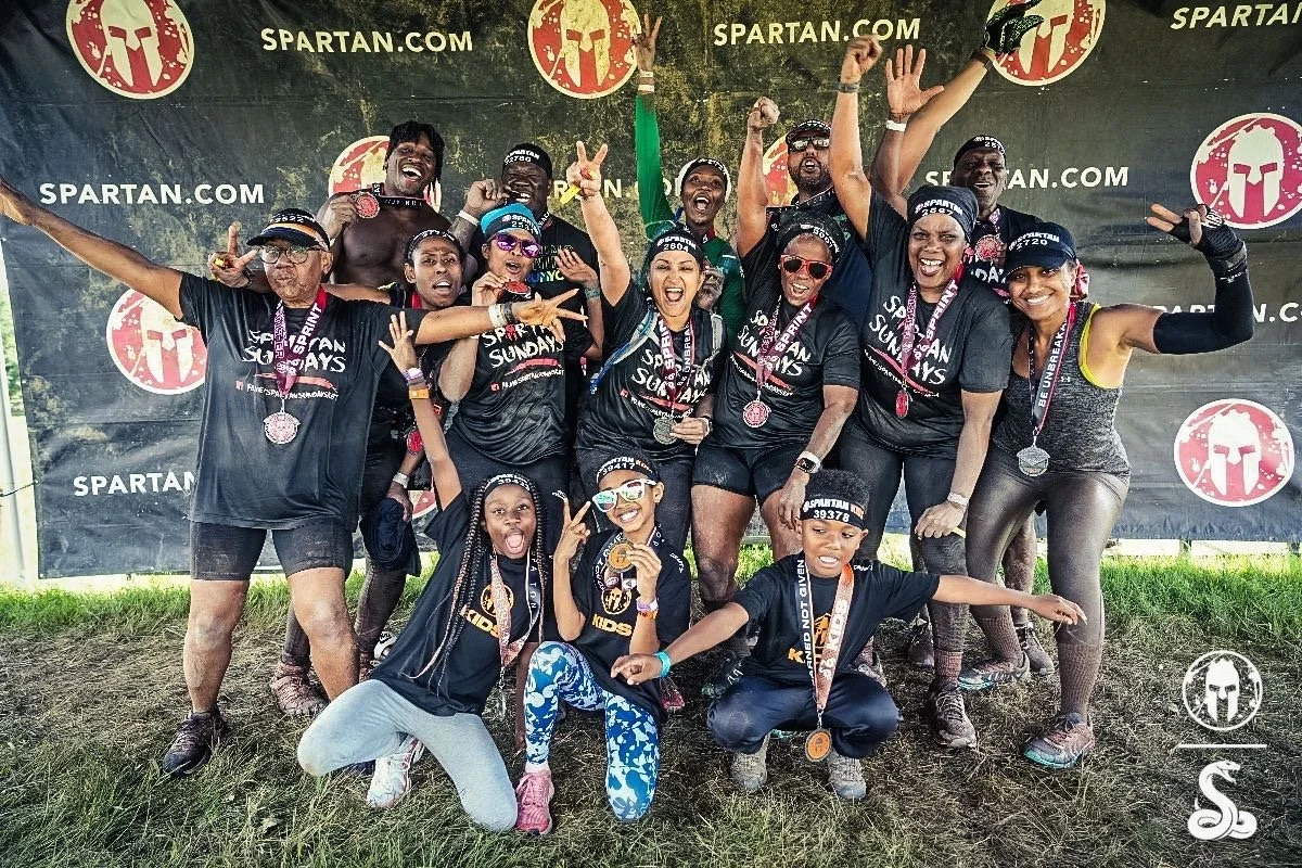 Group of people celebrating after a Spartan Race, wearing medals, black shirts with "Spartan Sundays," and headbands. They are posing energetically in front of a Spartan Race branded backdrop.
