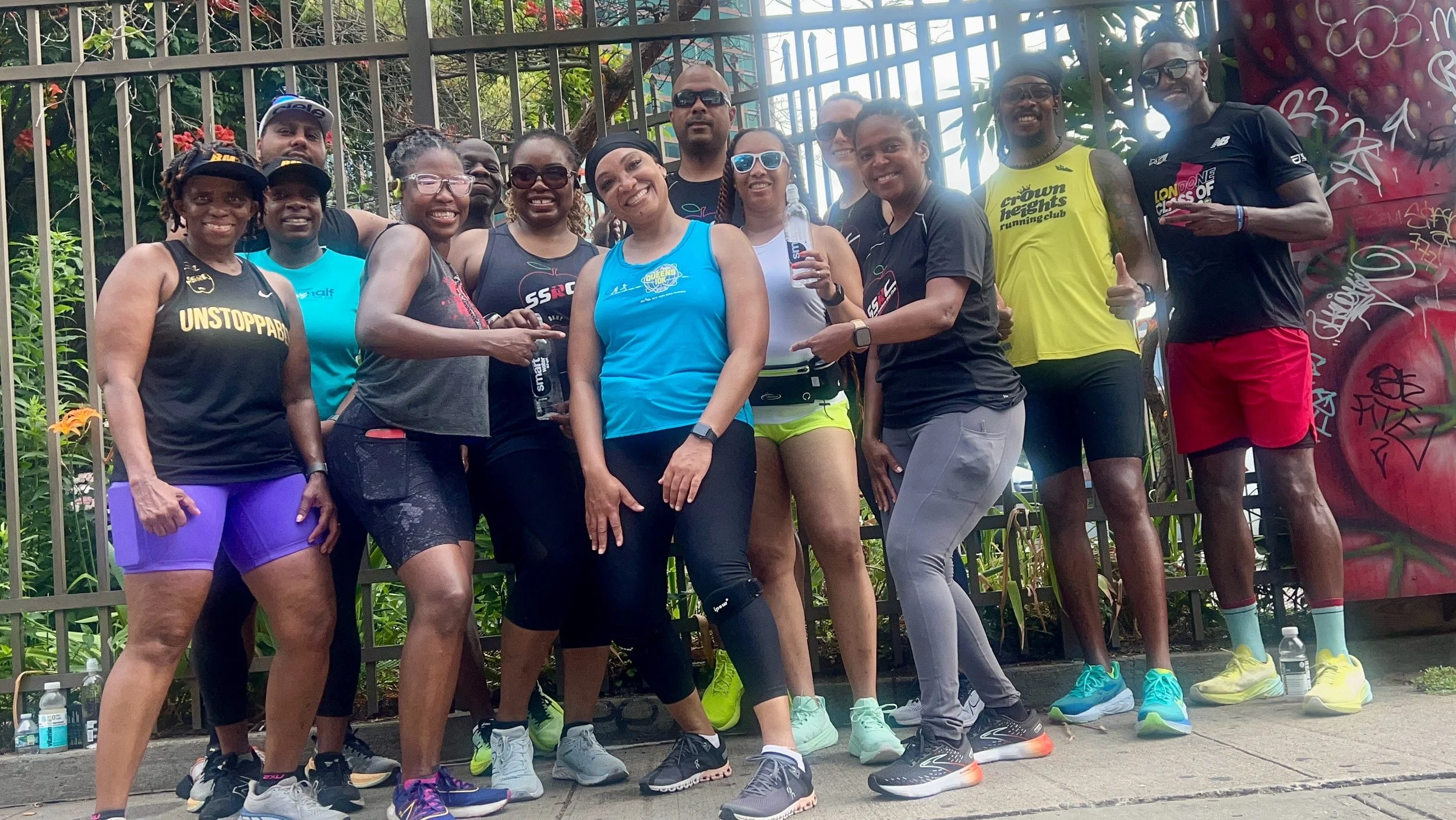 Group of people in athletic wear posing outdoors near a fence and graffiti wall.