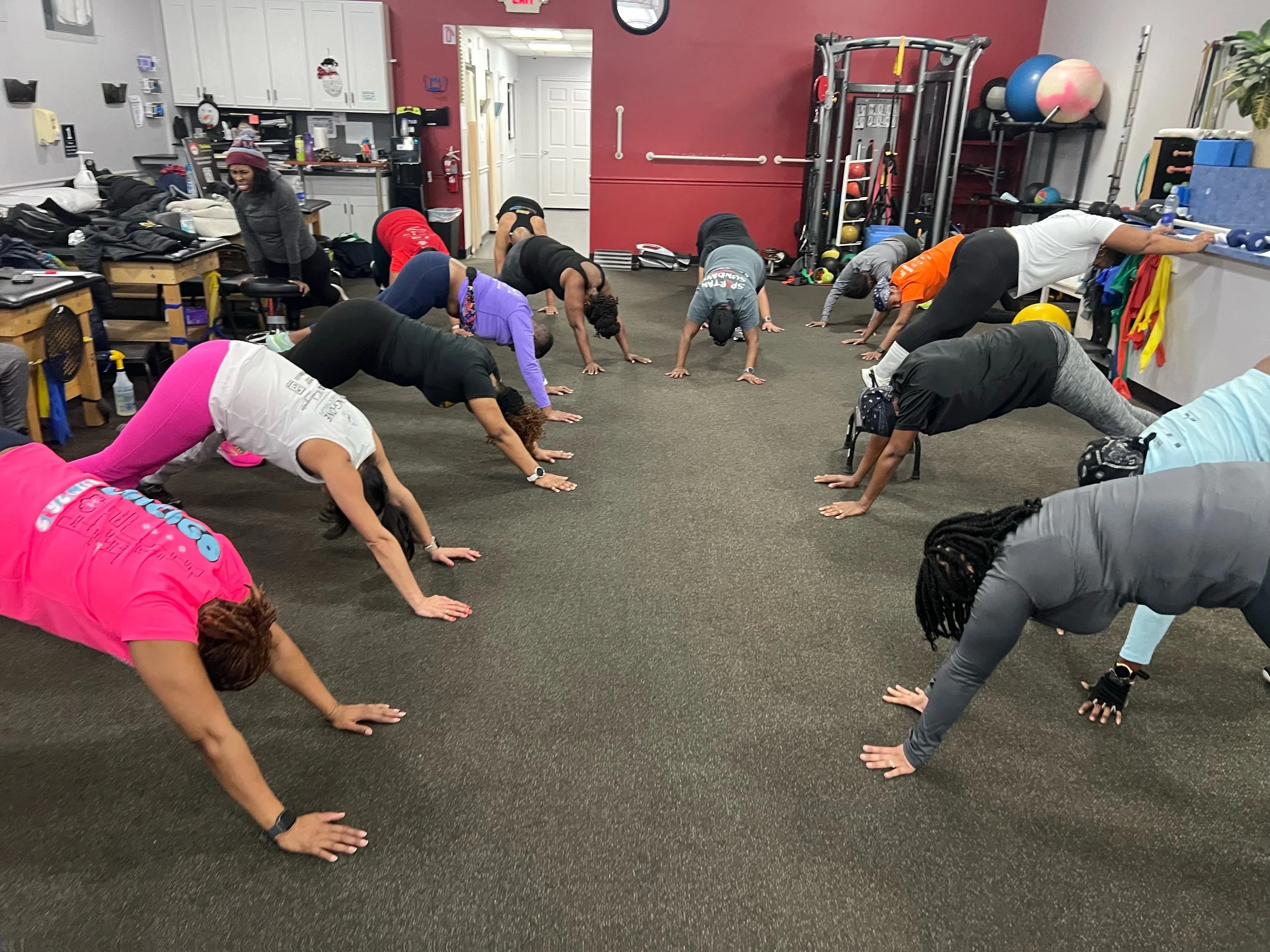 Group of people exercising in a gym, performing downward dog yoga pose on a carpeted floor, with workout equipment in the background.
