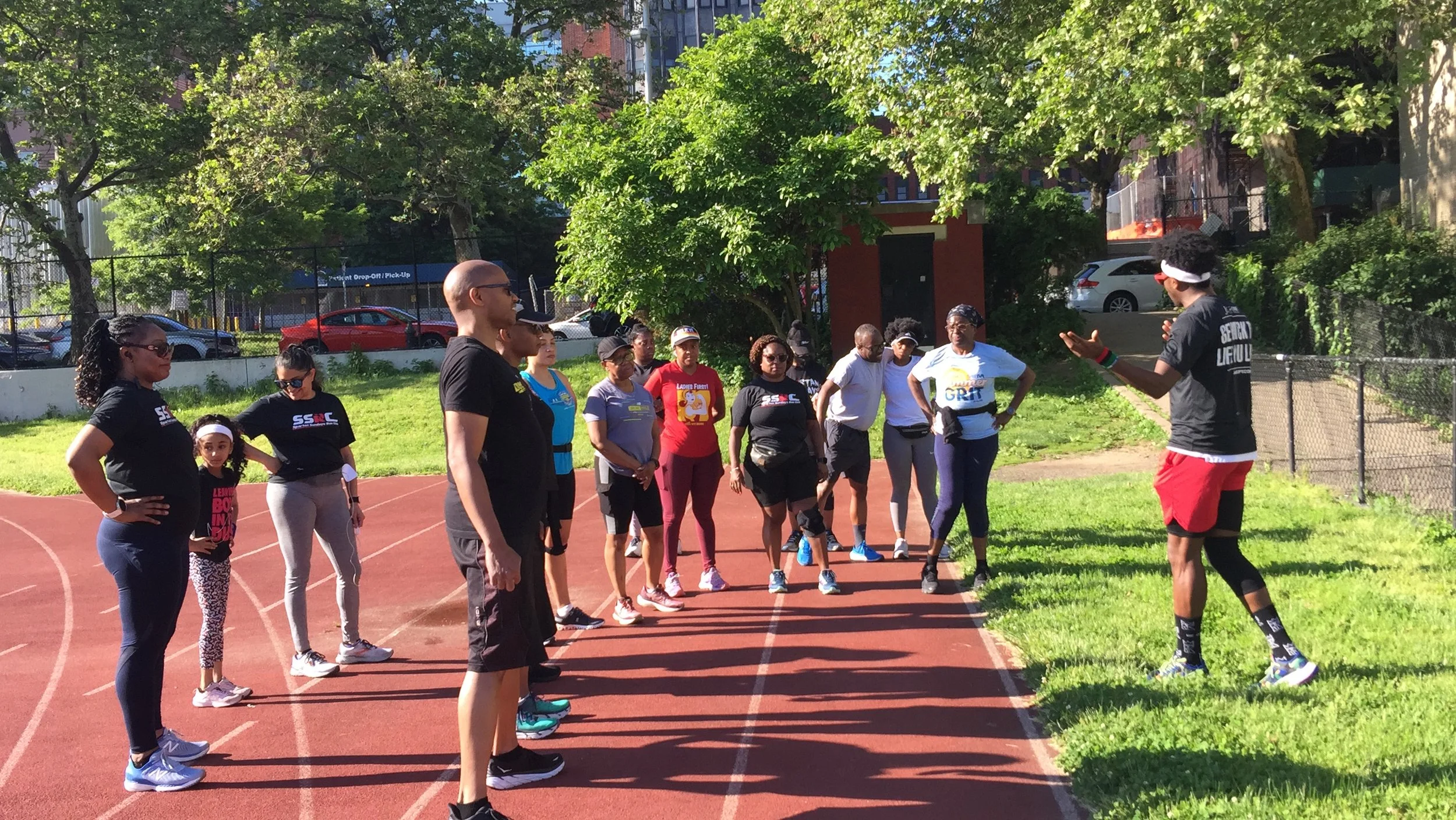 A group of people standing on a track in a park, dressed in athletic wear, facing an instructor. The instructor appears to be giving directions or instructions. Trees and grass are visible in the background.