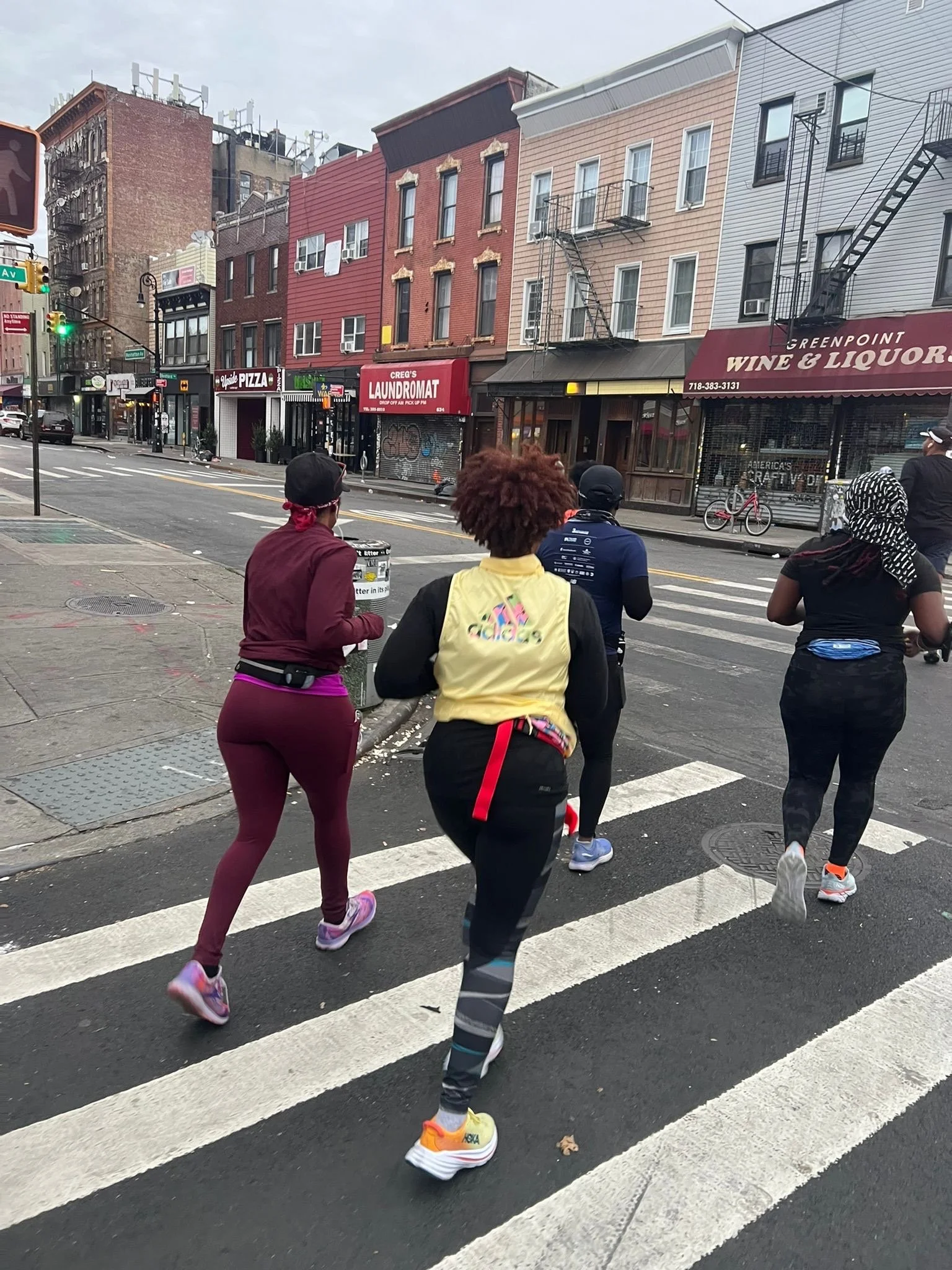 Four people jogging across an urban street crosswalk, passing by storefronts including a pizzeria, laundromat, and wine shop.