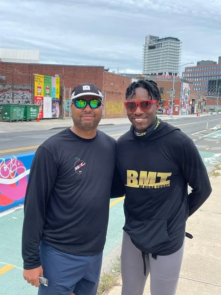 Two men standing on a city street with graffiti-covered brick buildings and urban art. They are wearing sunglasses and sportswear, smiling at the camera. A high-rise building is visible in the background.