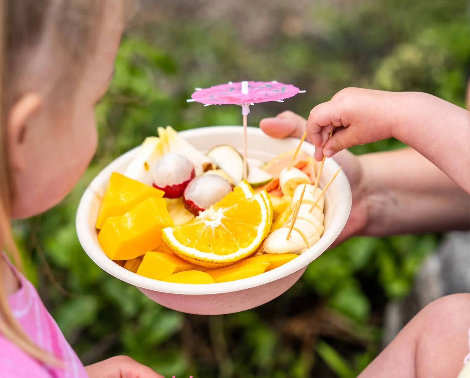 A child holding a bowl of assorted fruit, including orange slices, sliced bananas, mango, and lychee, with a pink paper umbrella.