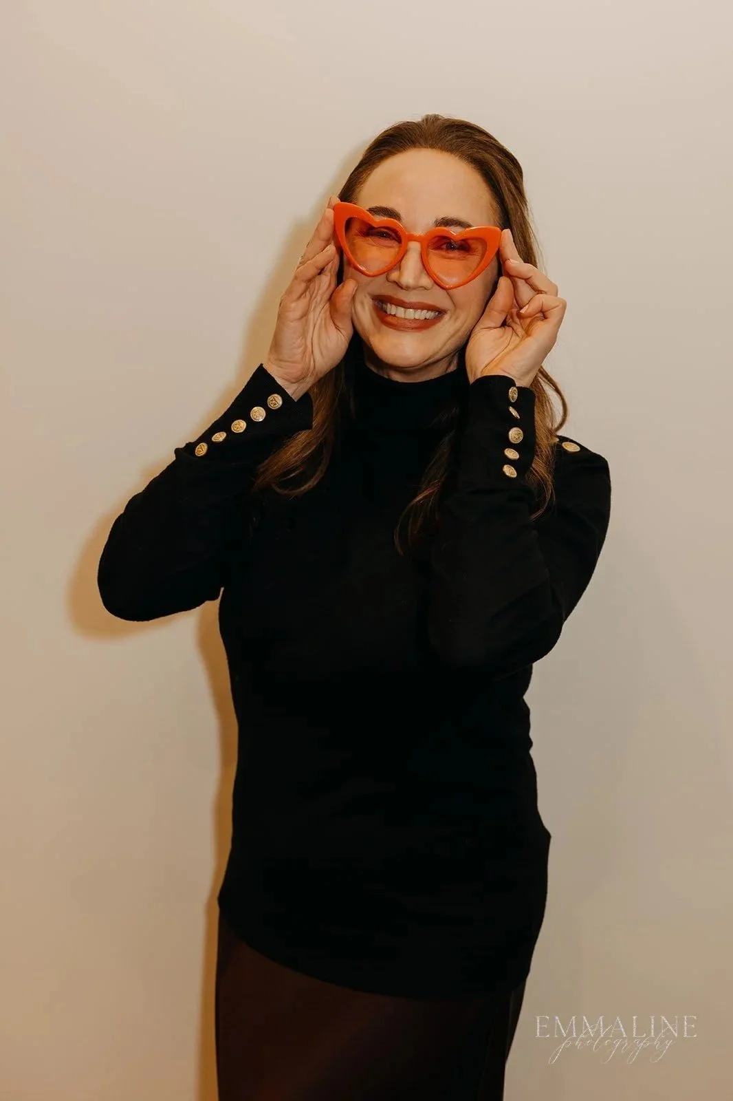 Woman smiling while wearing large orange heart-shaped sunglasses and a black outfit with gold buttons, standing against a plain wall.