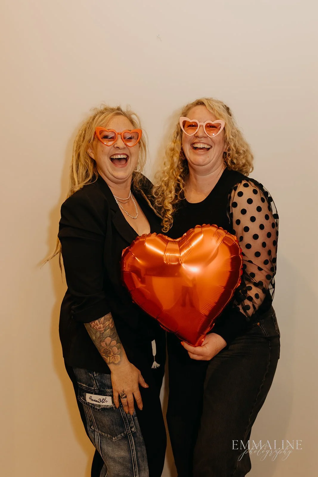 Two women laughing and holding a shiny red heart-shaped balloon, wearing colorful heart-shaped glasses, against a plain wall.