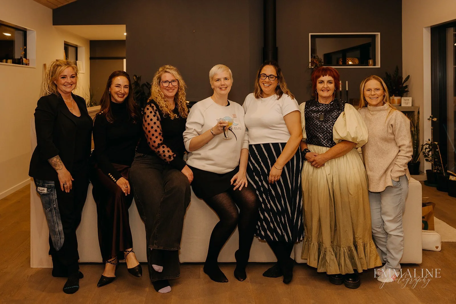 Group of eight women smiling, standing indoors against the back of a couch, dressed in casual and semi-formal attire.