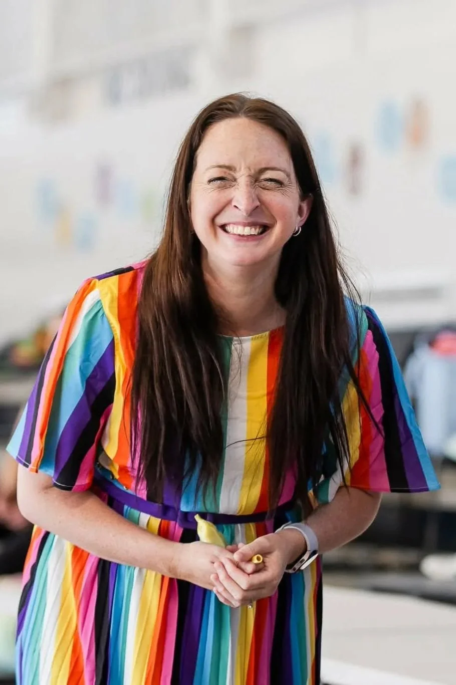 A cheerful woman with long brown hair wearing a rainbow-colored dress, smiling and holding a yellow pen, standing in an indoor setting.