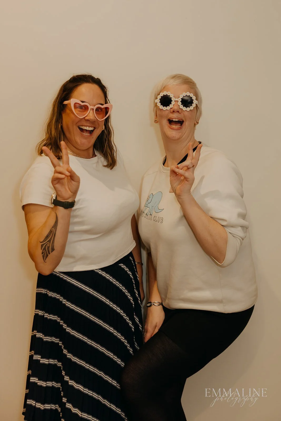 Two women smiling and posing with peace signs, wearing playful sunglasses, standing against a plain wall.