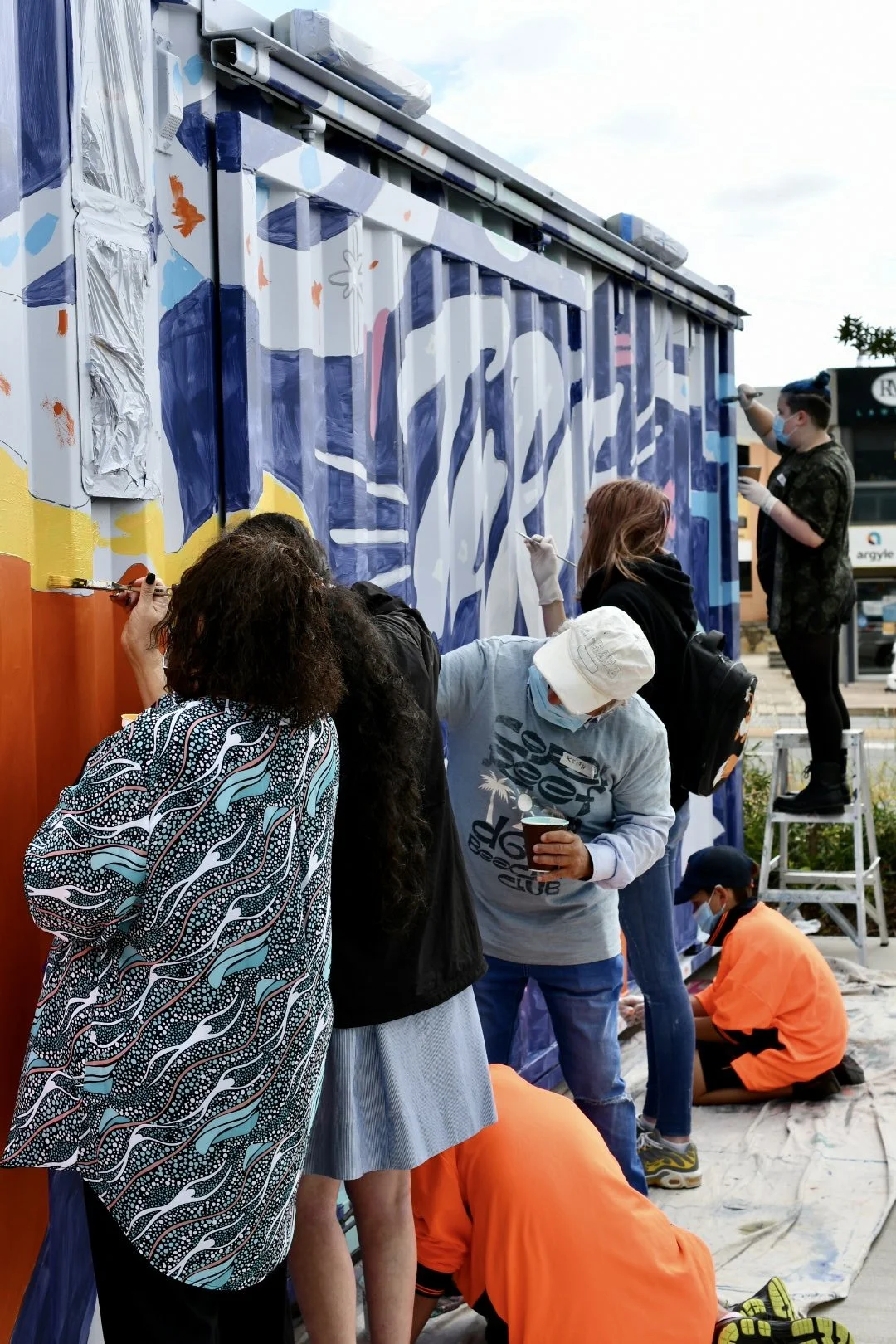 Community members of all ages and backgrounds painting a mural together, guided by artist Bohie Blackwood—capturing the spirit of co-design, creativity, and inclusive public art in action.