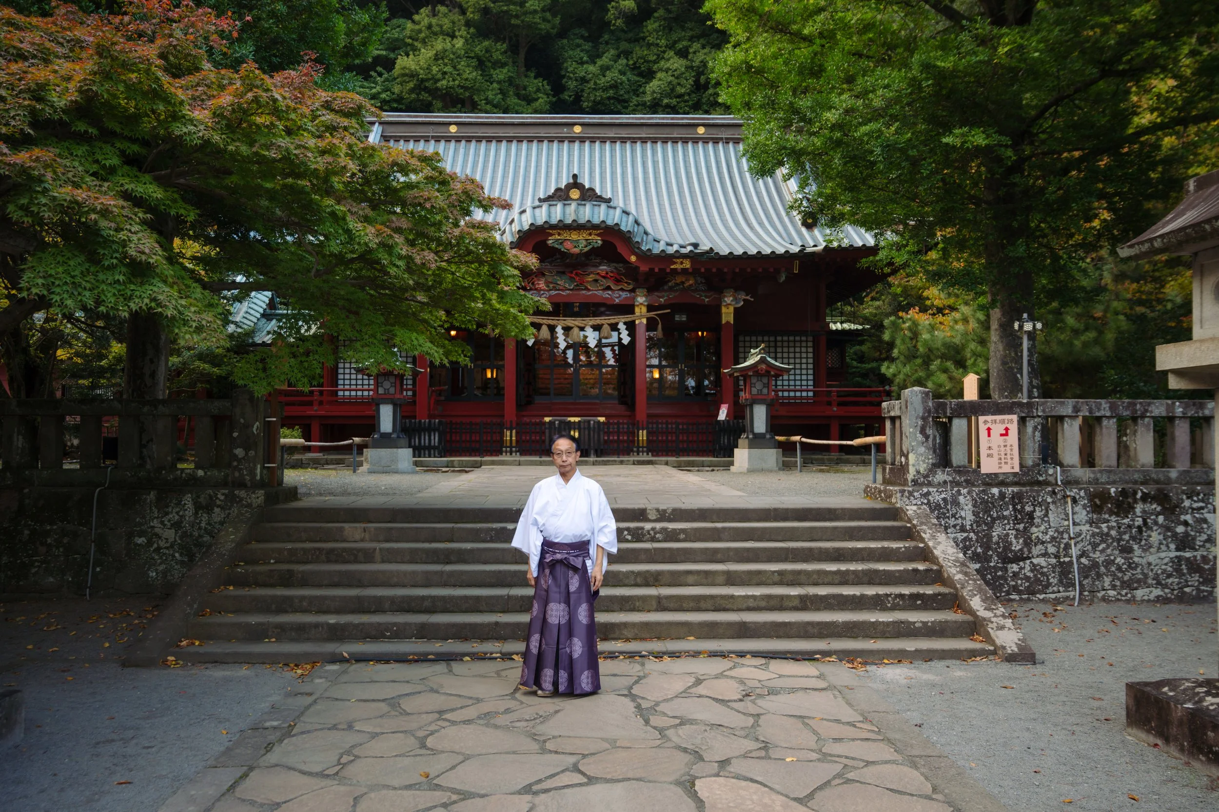 A person dressed in traditional Japanese attire standing on steps in front of a shrine, surrounded by lush green trees.