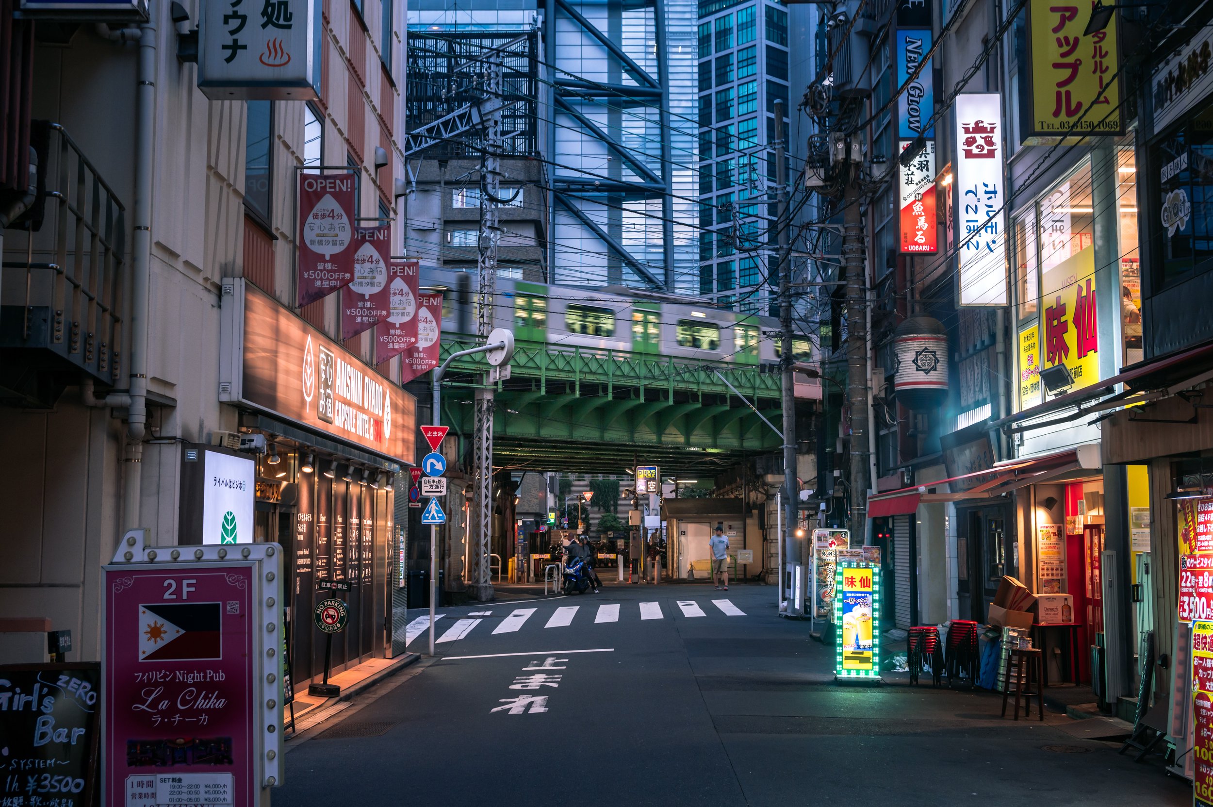 City street scene in Japan during evening, with buildings, signs in Japanese, a train crossing, and a green train passing overhead.