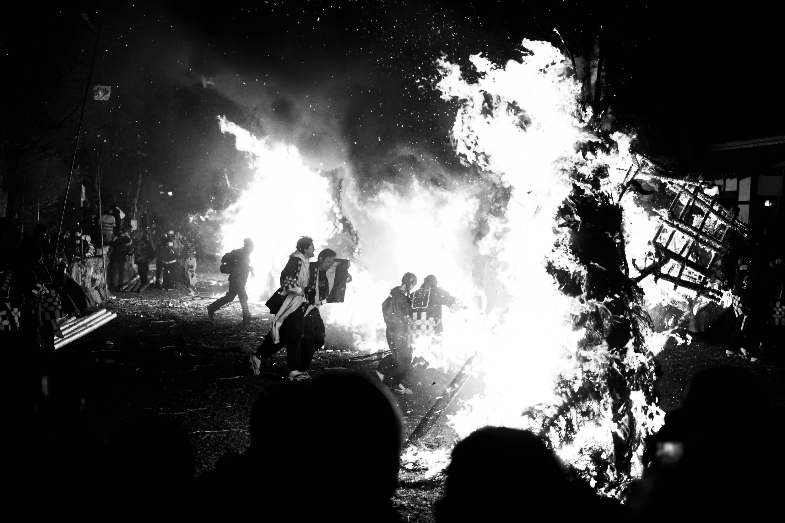 People walking away from large flames and fire debris at night in a chaotic scene.