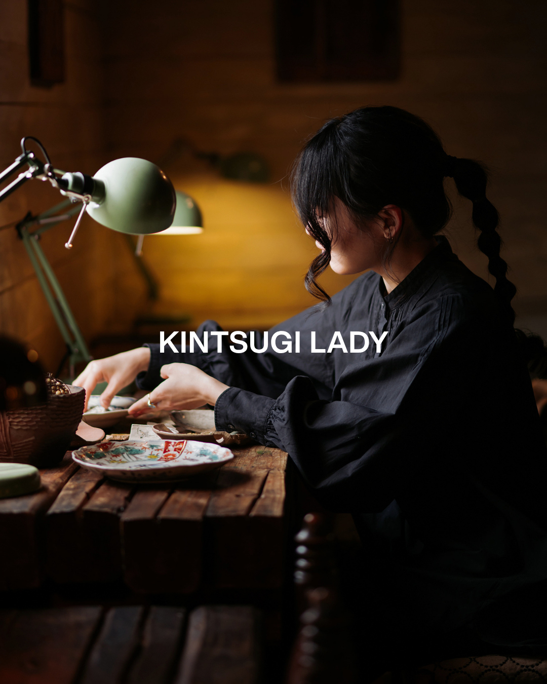 A woman with black hair in braids wearing a black shirt, sitting at a wooden desk with decorative plates and a basket, working under green desk lamps in a cozy, dimly lit room.