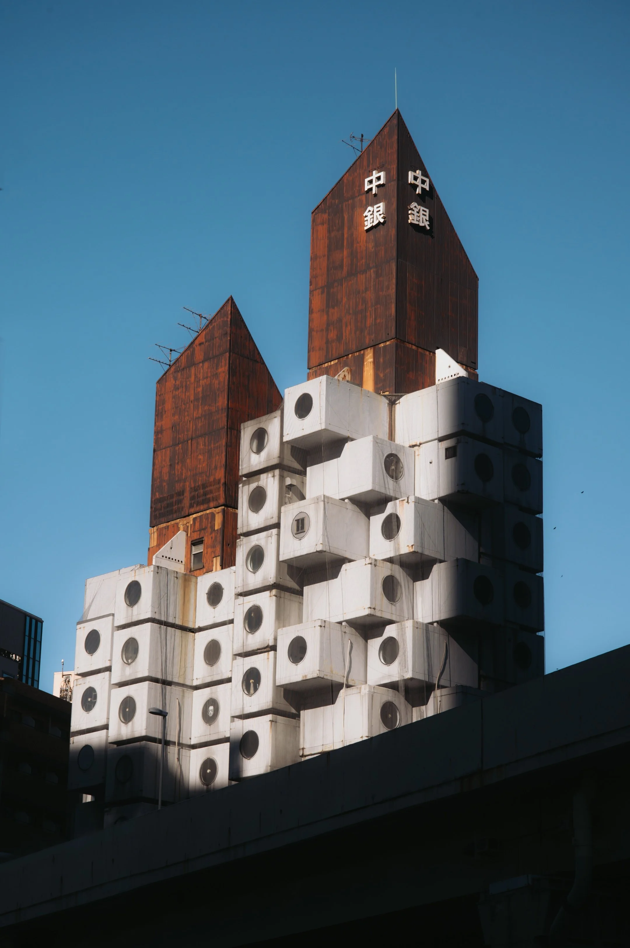 The image shows the top part of a modern building with a unique architectural design. The structure is made of stacked white blocks with circular windows and features two tall, rust-colored towers with some Chinese characters on them. The background 