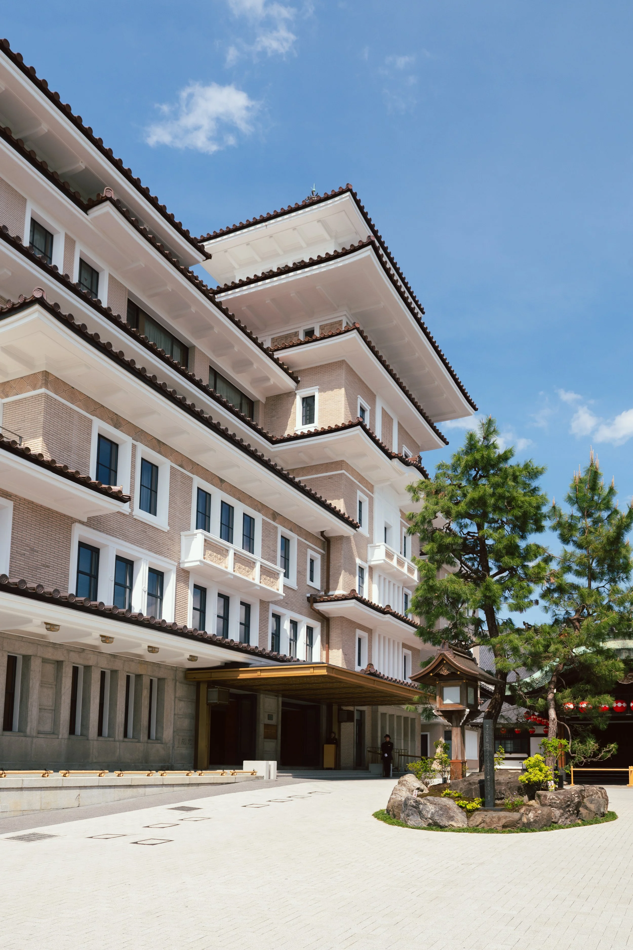 A modern multi-story building with traditional Asian architectural features, including a tiled roof and decorative elements, situated in a landscaped outdoor area with rocks, plants, and a trees under a clear blue sky.