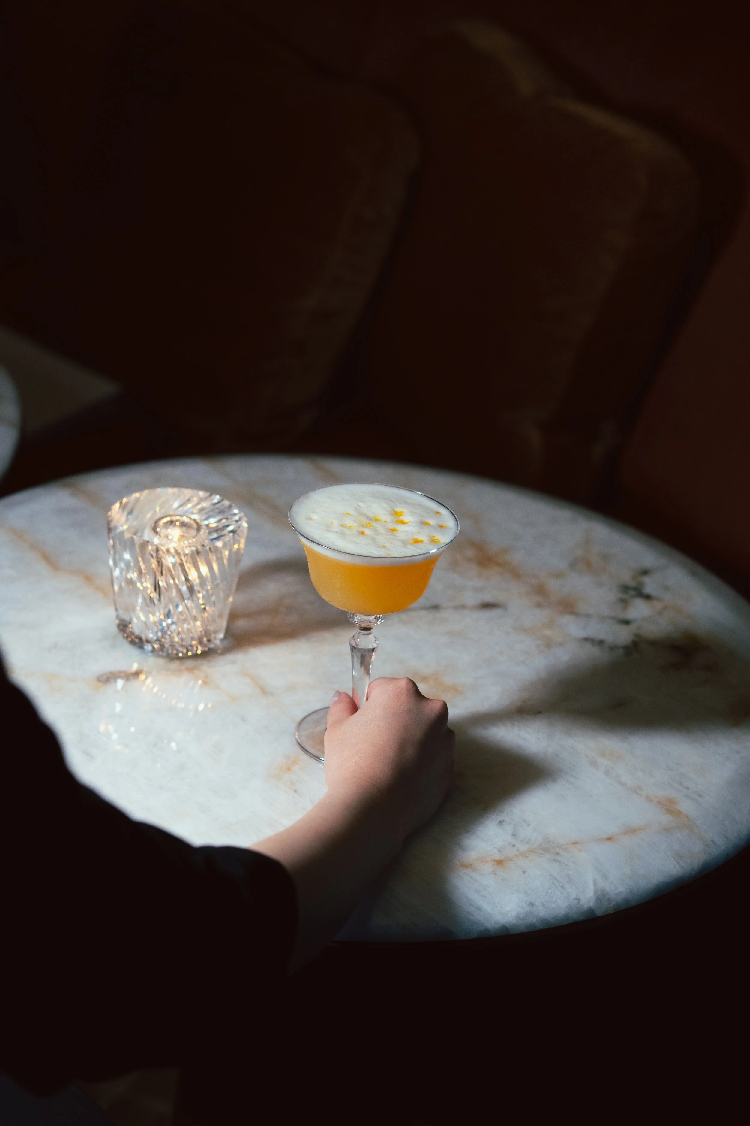 A hand holding a cocktail glass with a creamy drink topped with orange zest, on a marble table with a decorative glass candle holder in the background.