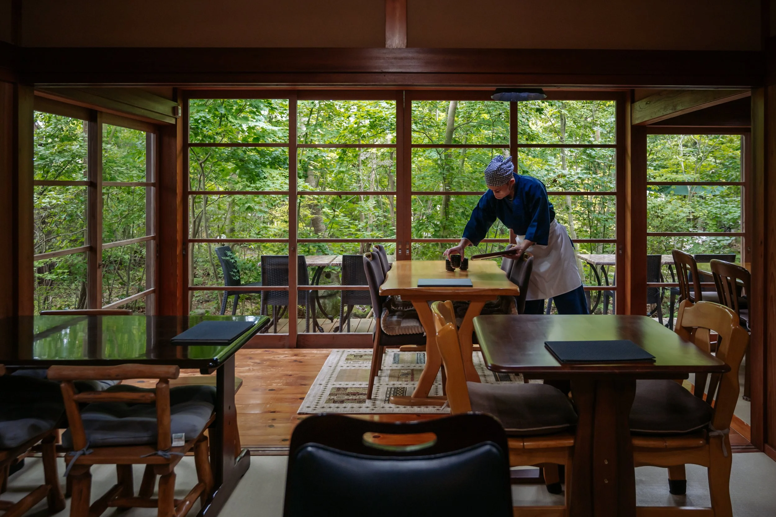 A person wearing traditional Japanese clothing and a headscarf bent over a wooden table, setting cups inside a room with large windows and a view of lush green trees outside.