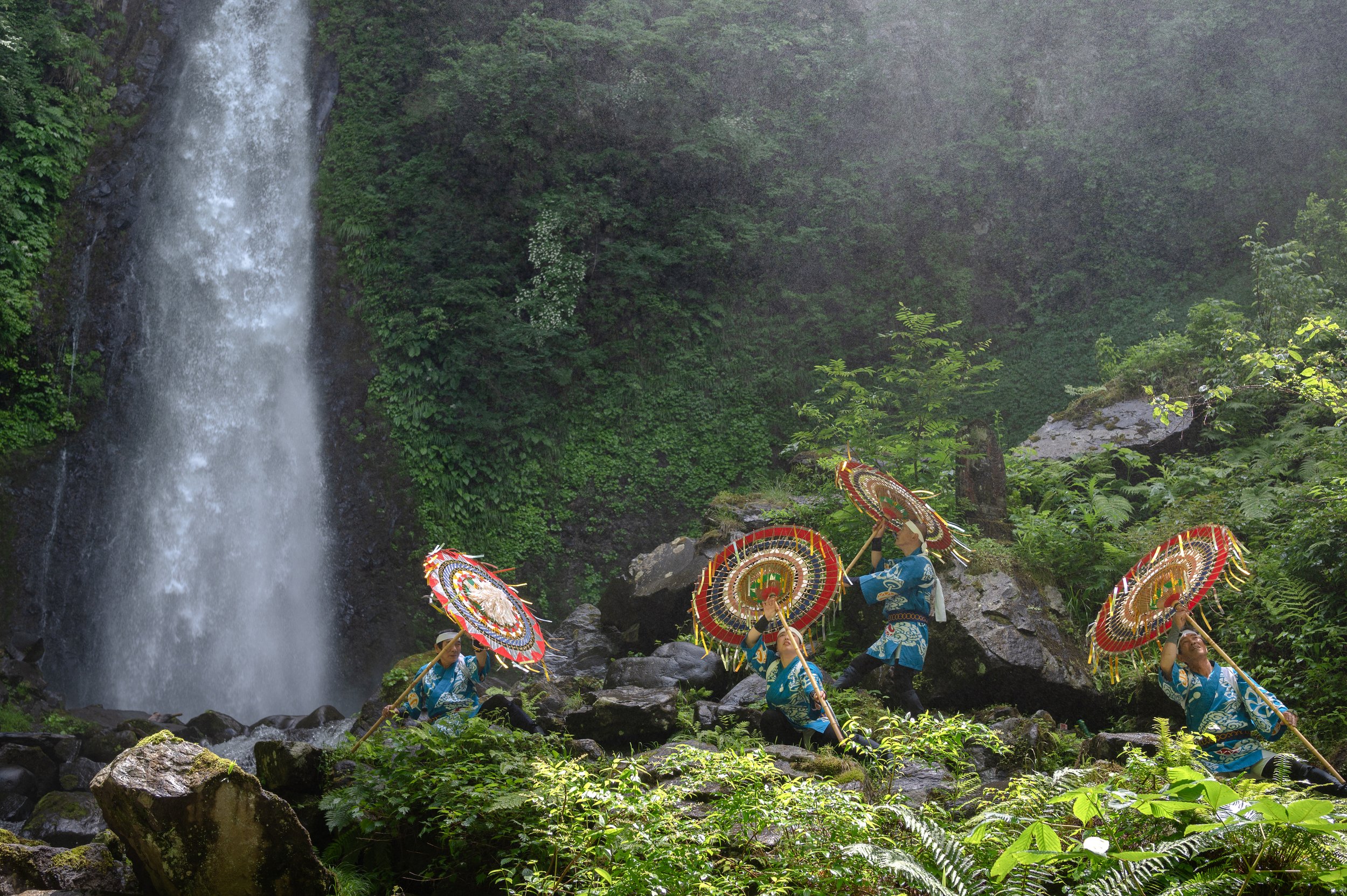 People dressed in traditional clothing holding umbrellas while standing near a waterfall in a lush green forest.
