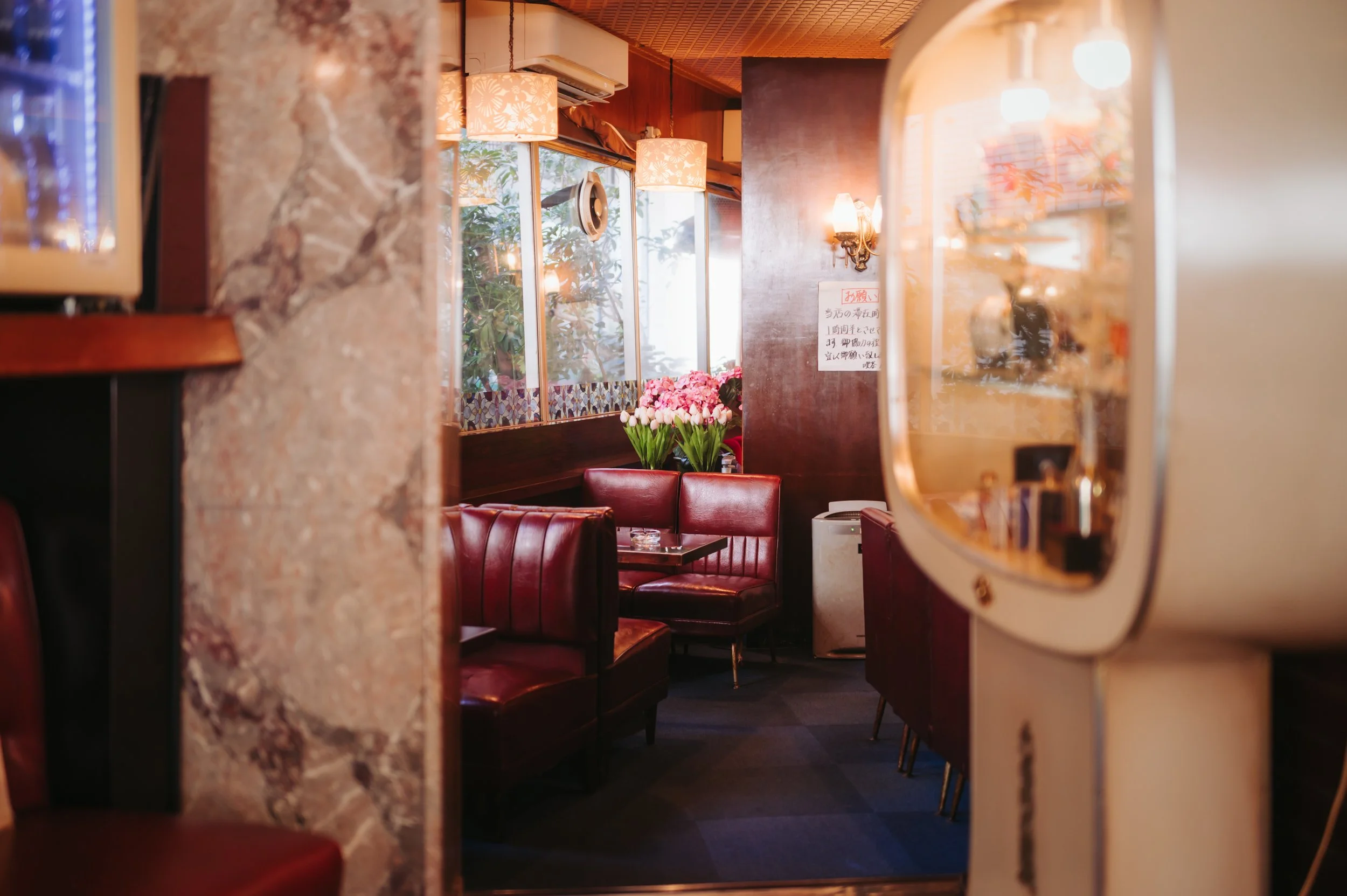 A cozy restaurant interior with red leather booths, a flower arrangement of pink tulips, and warm lighting, seen through a decorative mirror and a partial stone wall.