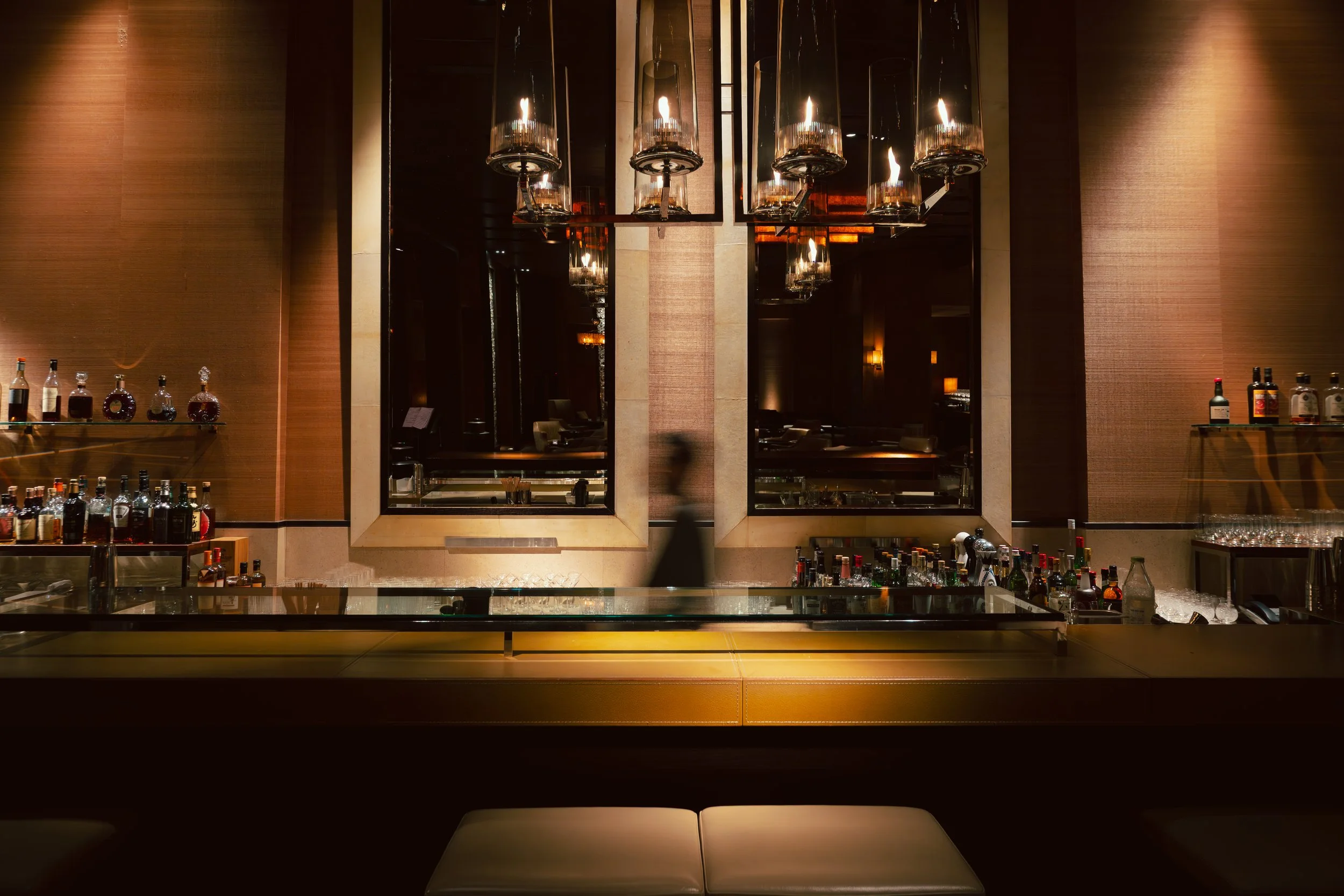 A modern bar with a sleek countertop, bottles of alcohol on display, large window mirrors, and hanging pendant lights illuminating a warm-toned interior.