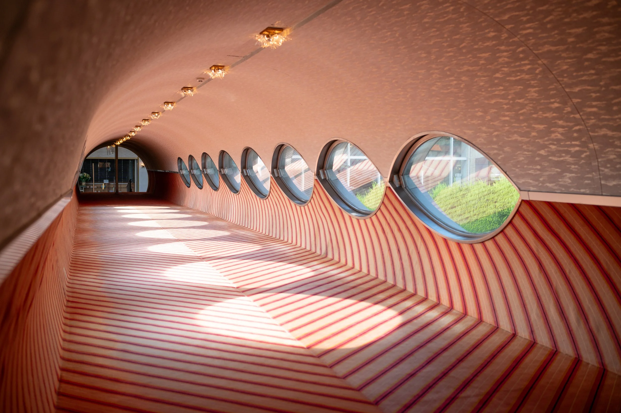 Interior view of a modern building corridor with red and beige striped walls and a curved design, large cylindrical windows on the right allowing natural light and views of greenery outside.