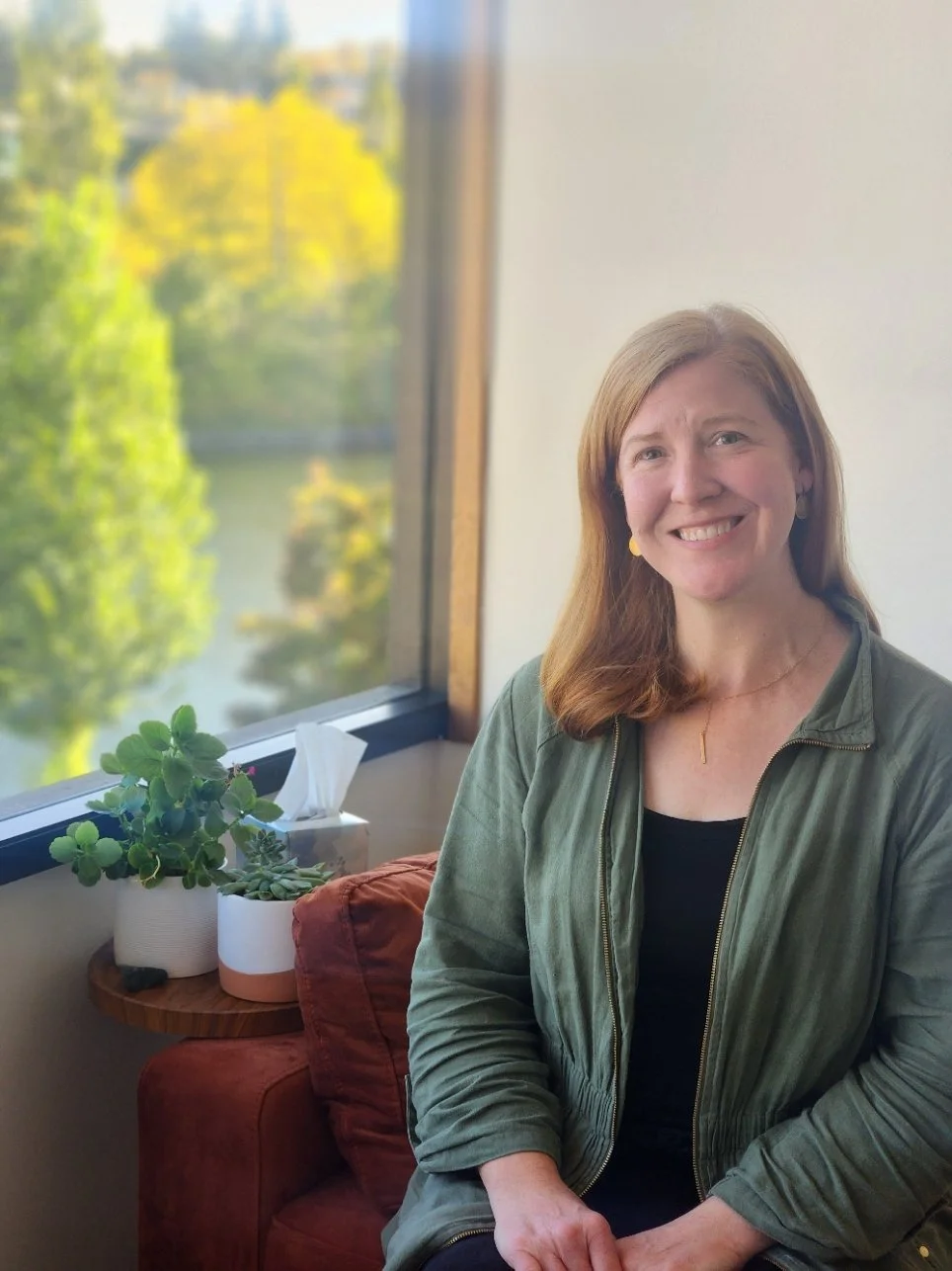 Kate, a Riverbank Therapy counselor, smiling in her Seattle therapy office with plants and a view of the water