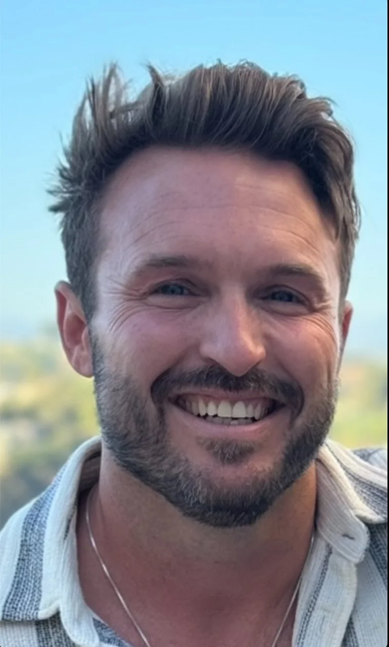 Close-up of a smiling low cost couples therapist in Seattle with short brown hair and a beard, wearing a collared shirt and a necklace, outdoors with a blurred blue sky and greenery in the background.