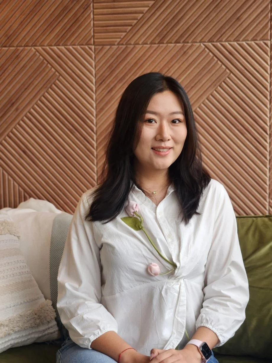 Seattle therapist Xinlei sits on a green couch with a brown textured wall behind her. She supports clients with depression, anxiety, trauma, chronic illness, and immigration issues.