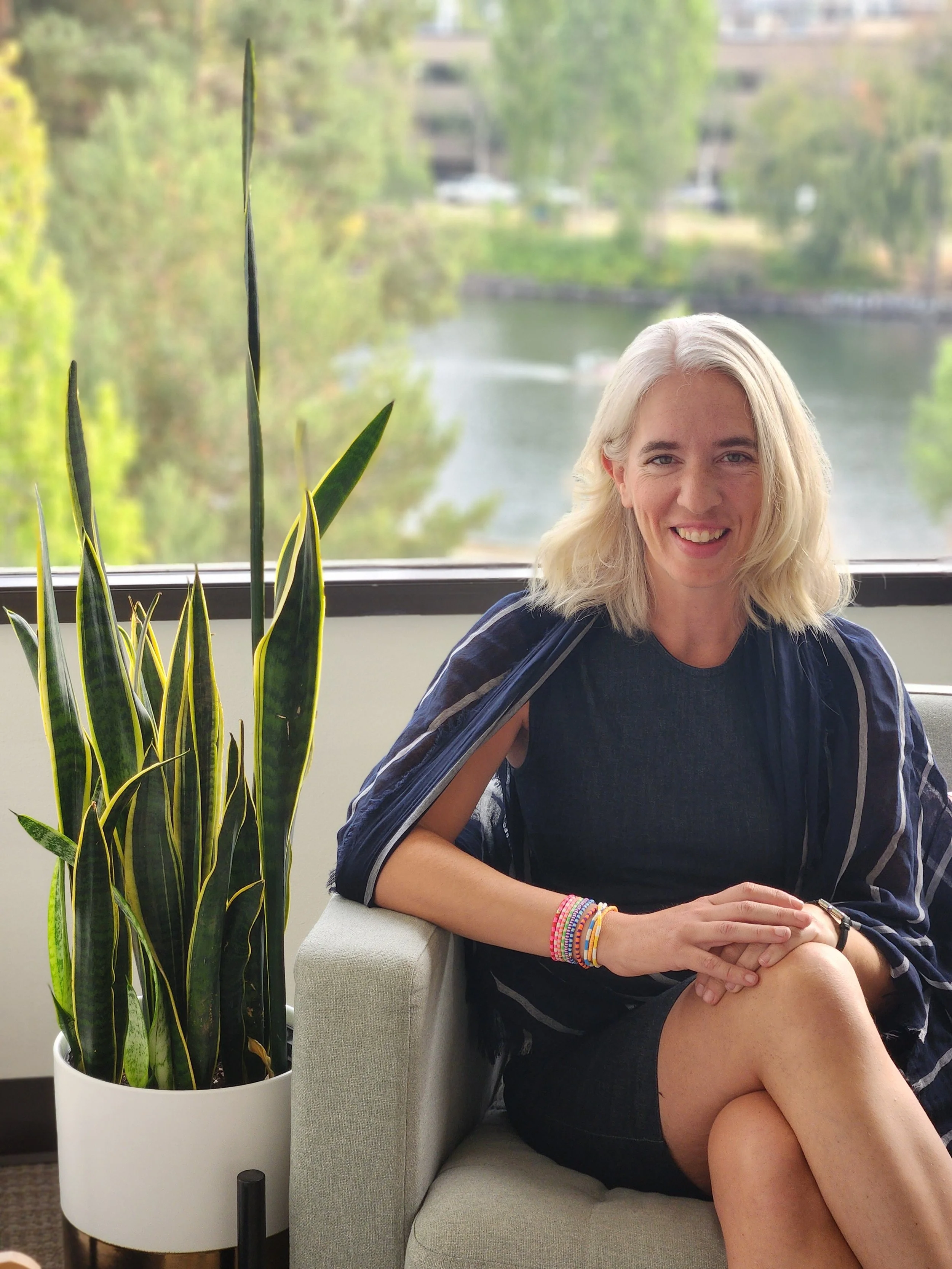 Seattle family therapist with blonde hair sitting on a couch next to a potted snake plant, outdoors with trees and a lake in the background.