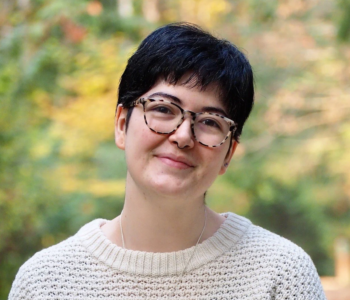 A therapist with short dark hair, wearing tortoiseshell glasses, a septum piercing, and a cream-colored knit sweater, smiling outdoors with blurred autumn foliage in the background.