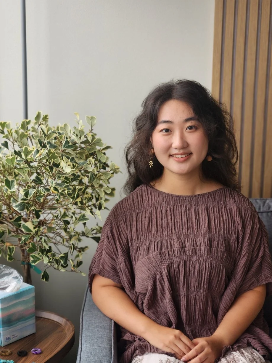 Seattle multicultural Seattle therapist sits in her office by a green tree. Nora provides therapy to immigrants, college students, teens, and those struggling with depression, anxiety and PTSD.