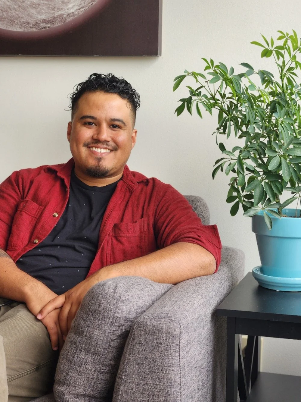 A smiling young man with short, curly black hair and facial hair, sitting on a gray armchair beside a large potted green plant with broad leaves in a blue pot, with a wall hanging behind him.