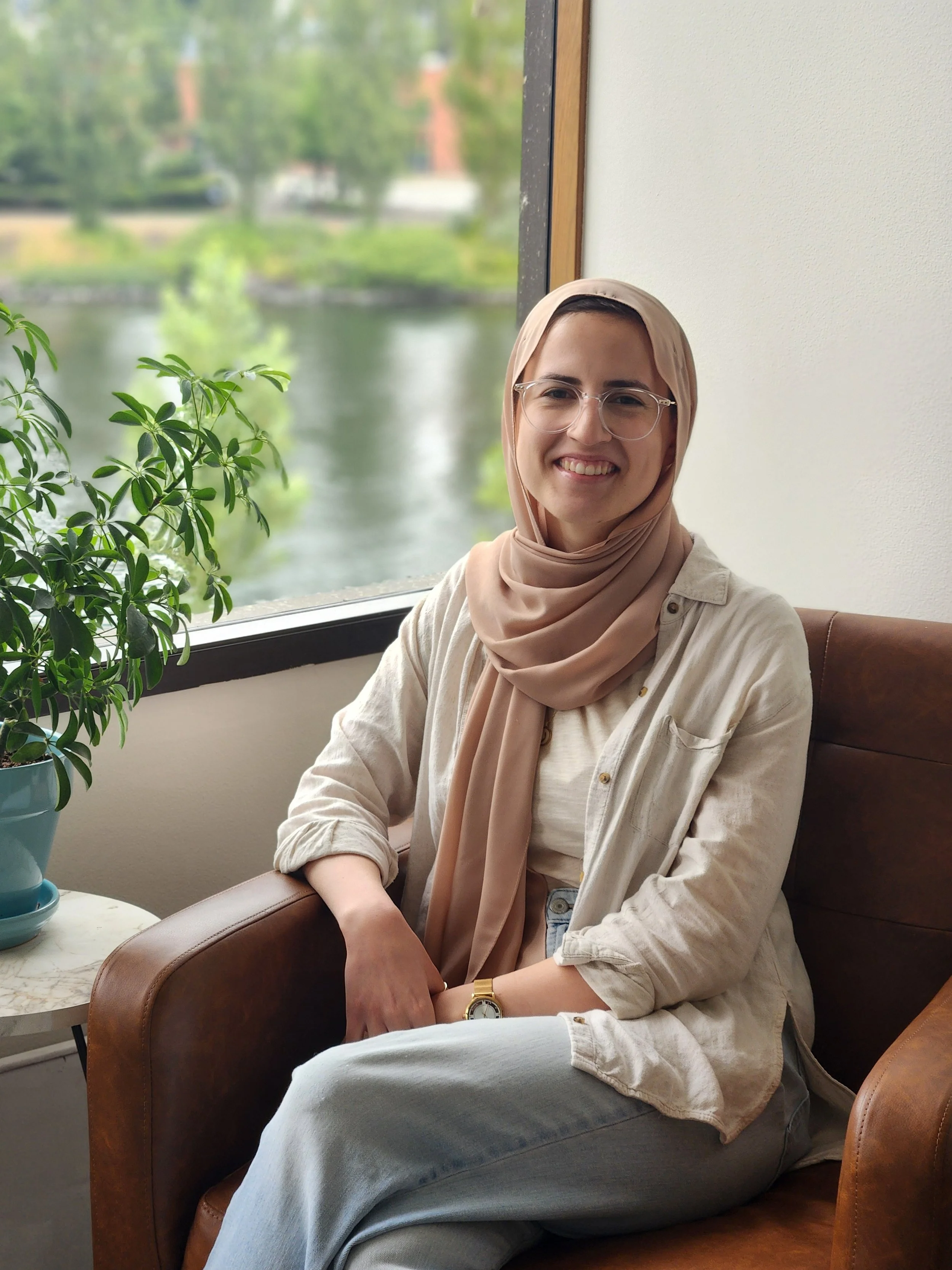 A smiling therapist with glasses and a beige hijab sitting in a brown leather armchair near a window with a view of trees and water outside, in queen anne, Seattle.