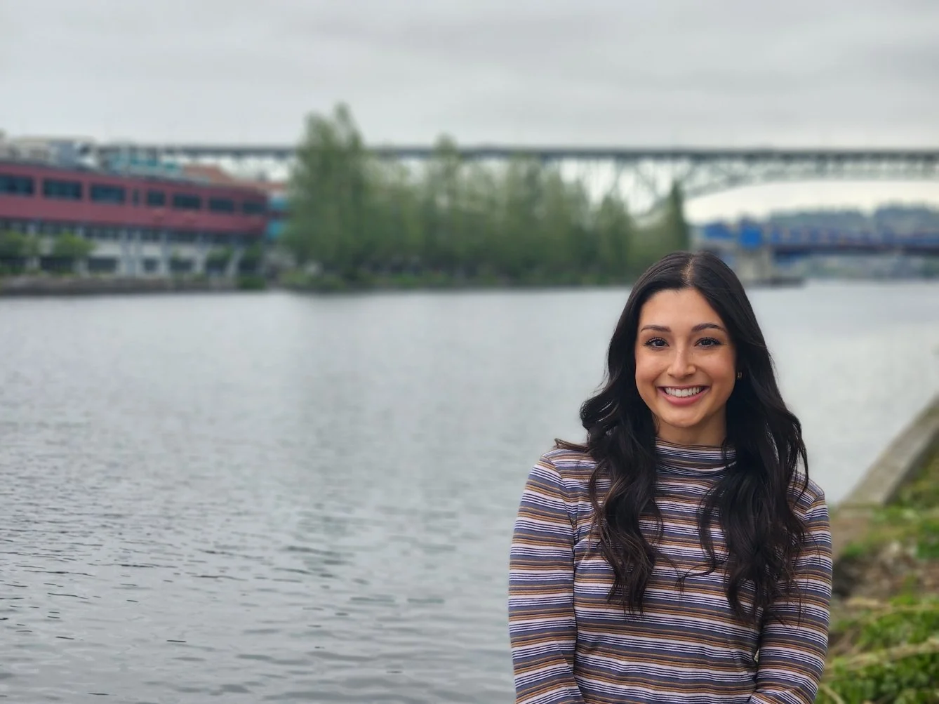 Seattle couples and trauma therapist stand outside on a cloudy day, by the Fremont canal and Aurora bridge. She has long dark hair and is smiling.