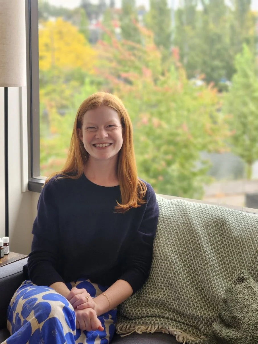 Raina, a Seattle couples and family therapist, smiles from a couch with a green woven blanket, in front of a large window looking out on fall trees.