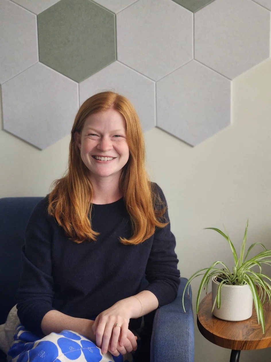 Raina, a Seattle couples therapist with long red hair, smiles while sitting in a blue armchair beside a spider plant in a cozy therapy office.