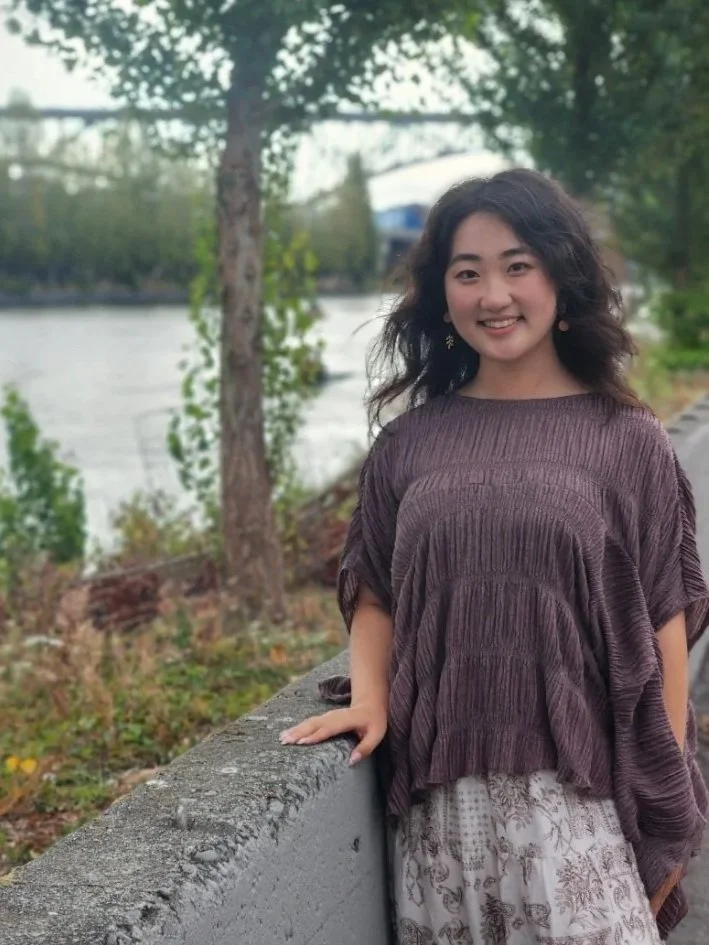 Seattle therapist Yinuo Wang stands outside by the Fremont canal, the water and trees are behind her. Nora specializes in working with immigrants, anxiety, depression, and PTSD.