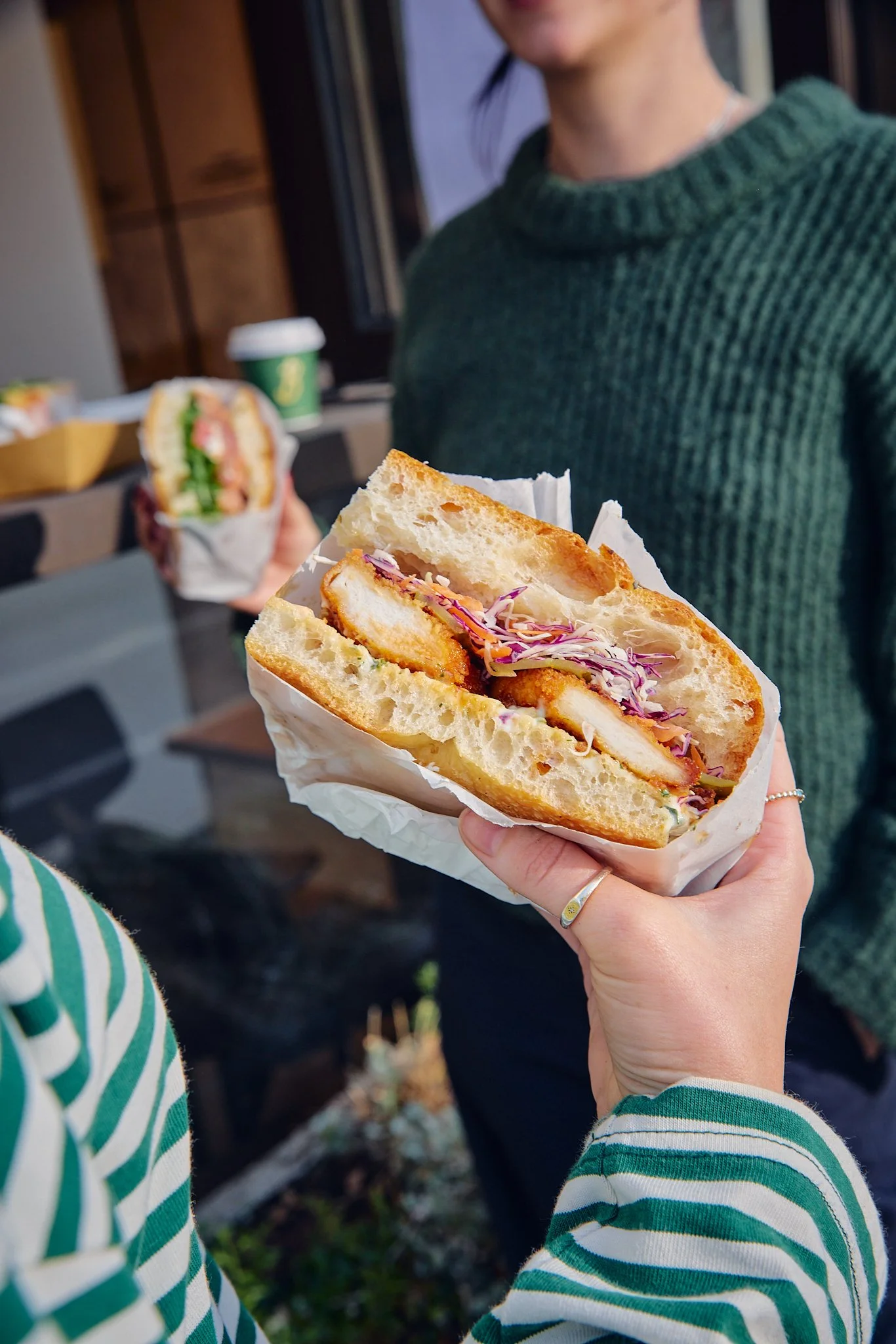 Close-up of a sandwich with fried chicken, coleslaw, and bread in hand, with a woman wearing a green sweater and another person with a striped sweater holding a second sandwich in the background.