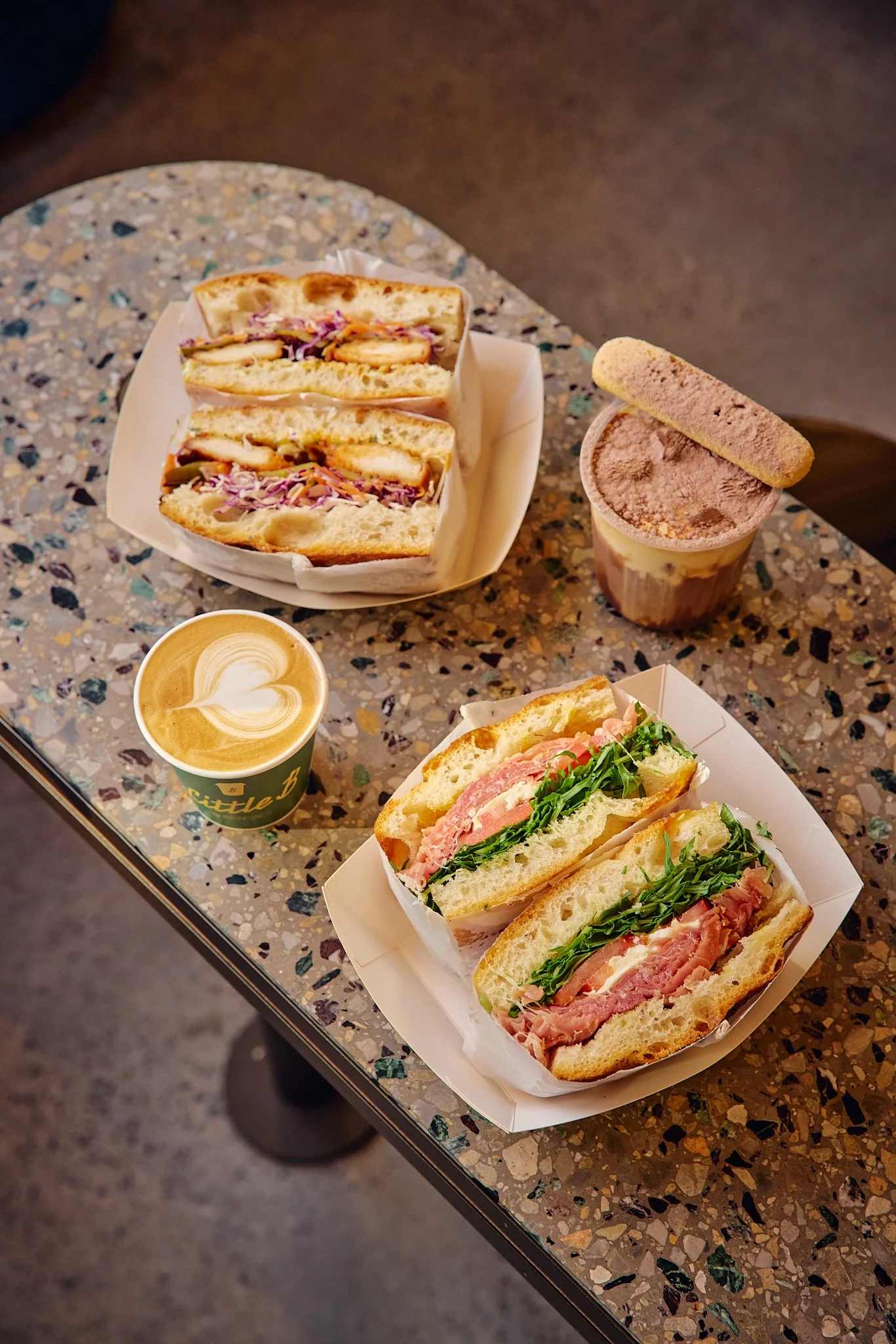 A terrazzo table with a sandwich, a cappuccino, and a coffee drink topped with a biscotti and powdered cocoa. The sandwich contains ham, tomatoes, arugula, and cheese.
