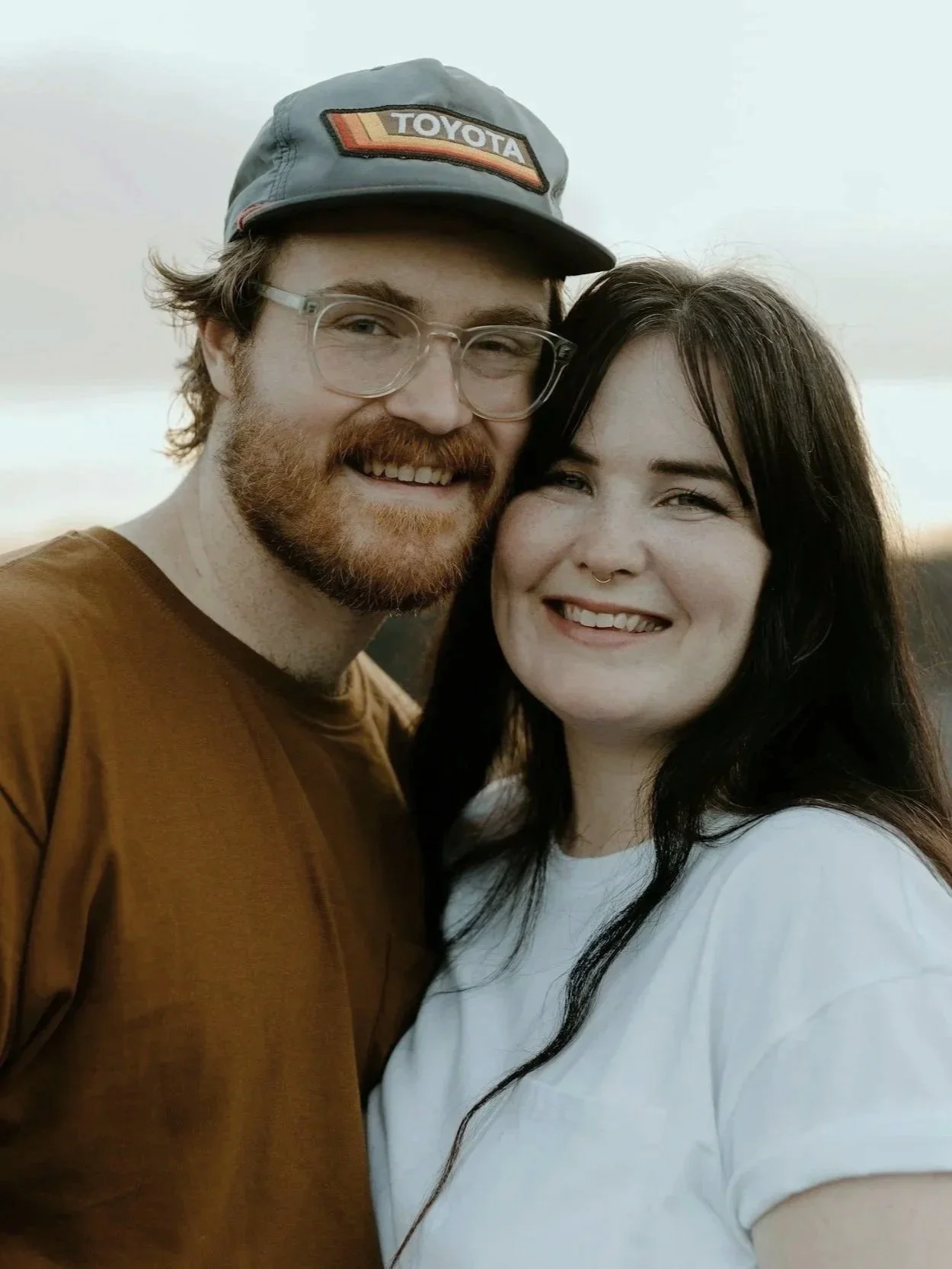 A smiling man with glasses, a beard, and a baseball cap standing close to a smiling woman with dark hair and a septum piercing, outdoors during sunset.