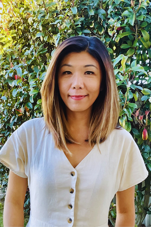 A woman with shoulder-length hair, wearing a white button-up shirt, standing outdoors in front of green leafy bushes.