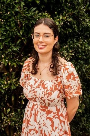 Young woman with glasses and wavy brown hair, smiling, standing outdoors in front of green foliage, wearing a floral dress with puffed sleeves.