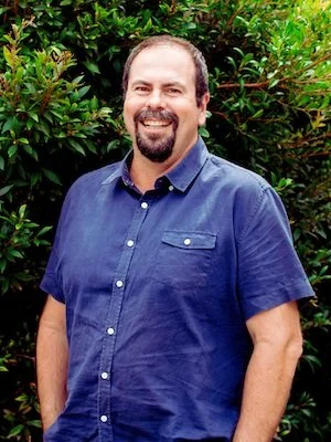 A man with dark hair, beard, and mustache smiling, wearing a blue short-sleeved button-up shirt, standing outdoors in front of green foliage.