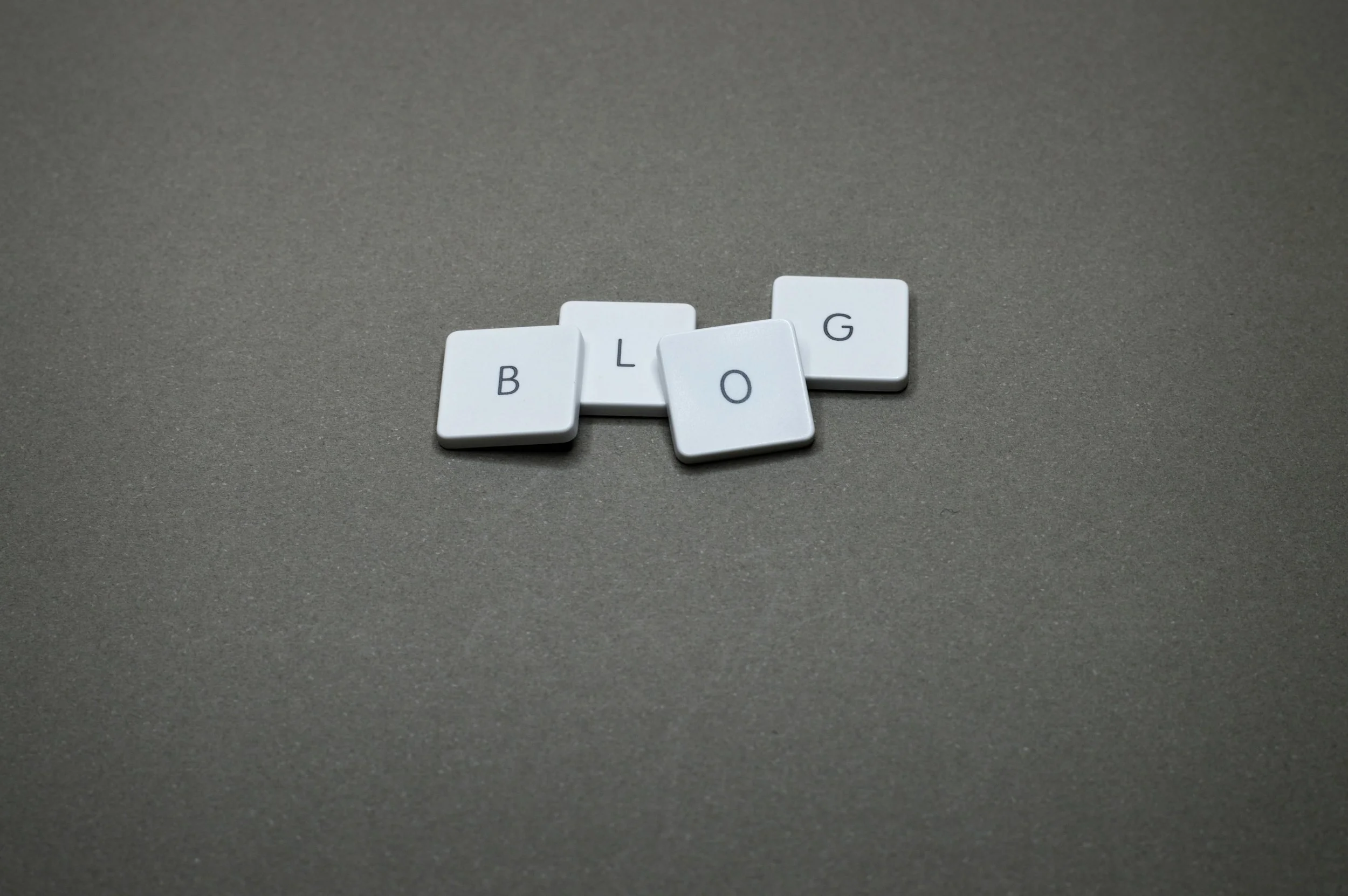 Four white tiles with black letters spelling out 'BLOG' are arranged on a gray surface.