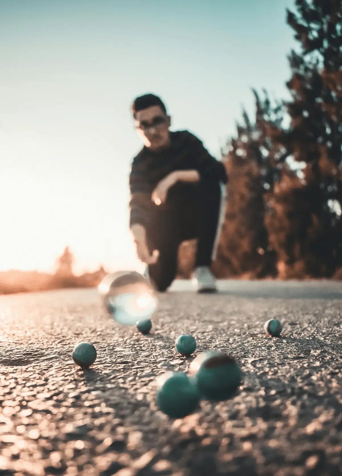 An adult kneeling on concrete, carefully observing a glass shooter marble as it rolls toward target marbles during an outdoor game.